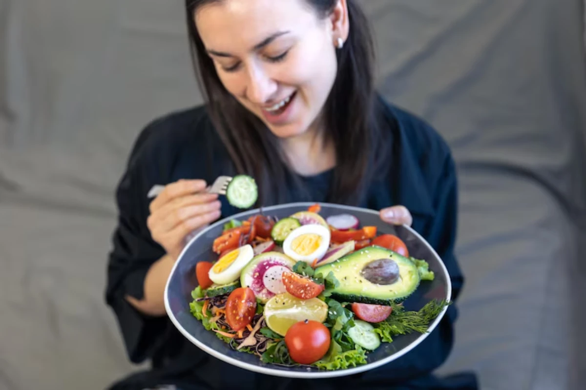 A person holds a large plate of colorful salad with avocado, cherry tomatoes, cucumber, and boiled eggs, set against a gray background.
