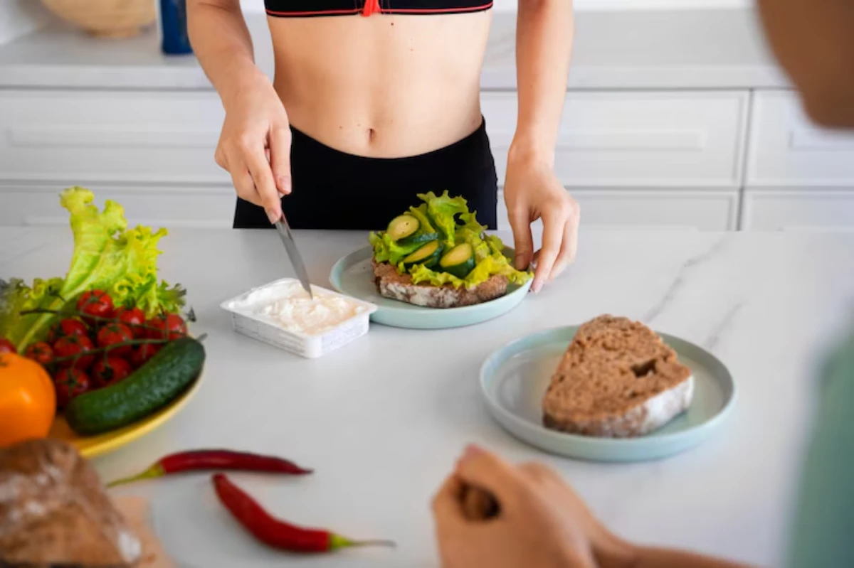 A person in activewear prepares a healthy meal with lettuce and cucumbers on whole grain bread, surrounded by fresh vegetables.