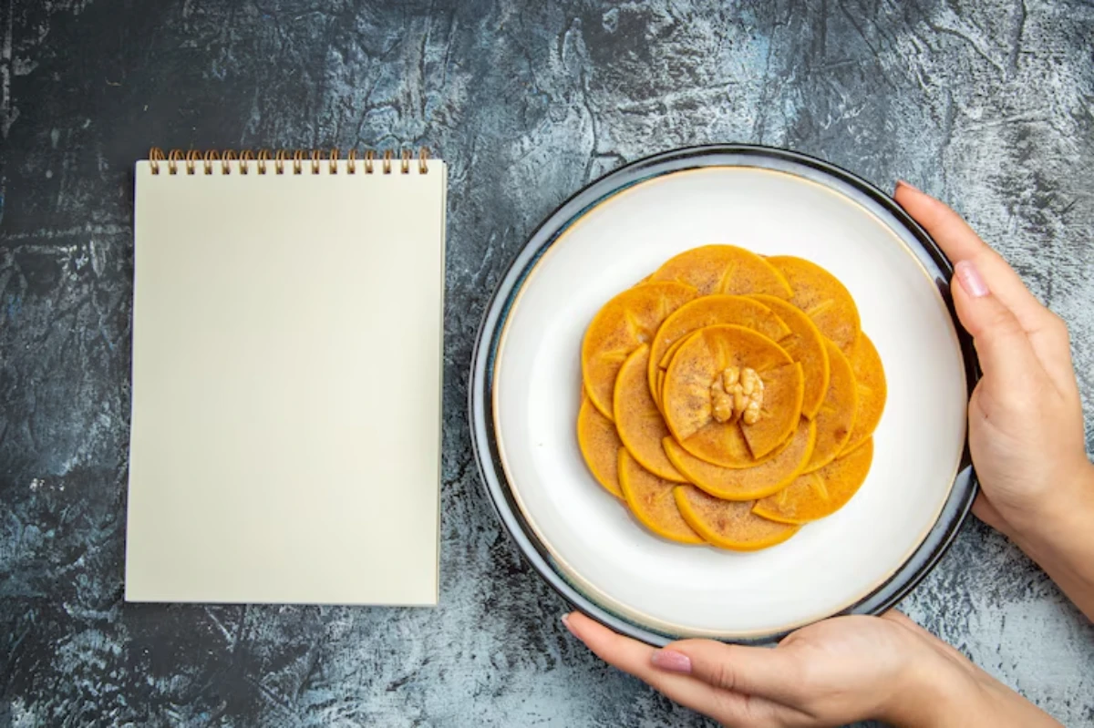 A plate of beautifully arranged orange dessert resembling a flower, with a walnut on top, beside a blank notepad.