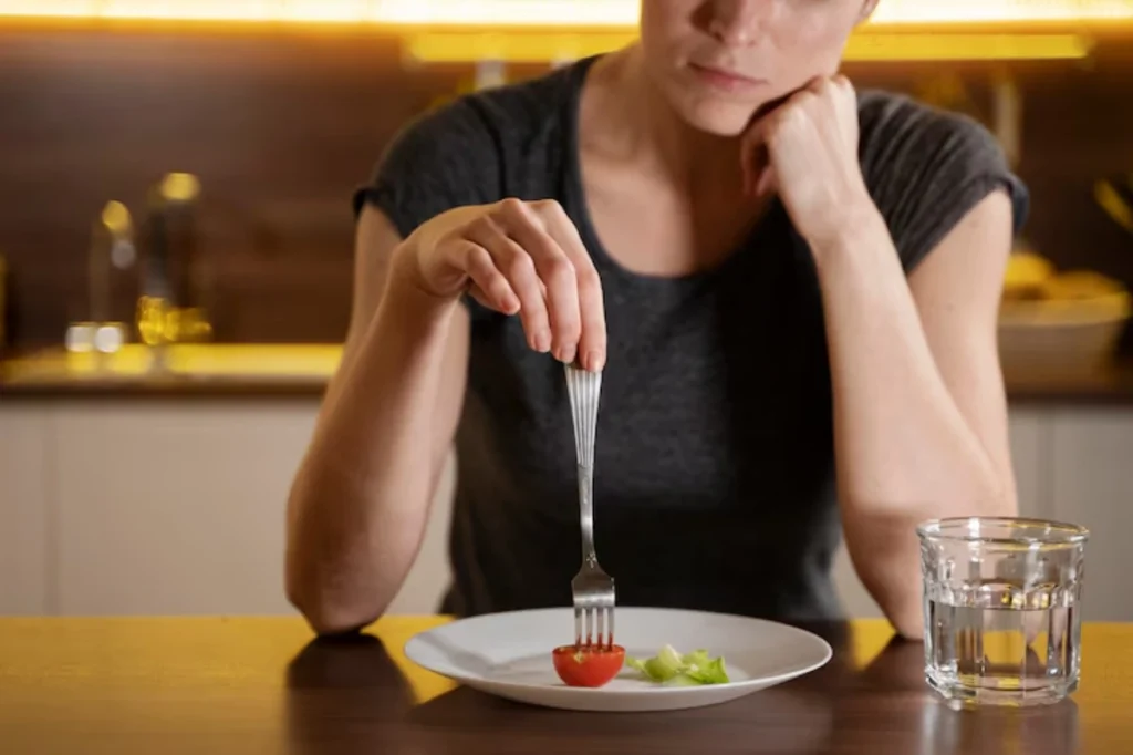 A woman in a grey shirt thoughtfully holds a fork piercing a cherry tomato on a minimal plate, contemplating a small meal.