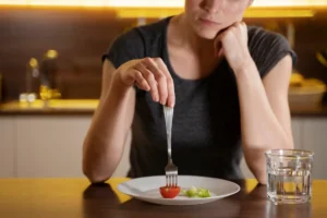 A woman in a grey shirt thoughtfully holds a fork piercing a cherry tomato on a minimal plate, contemplating a small meal.