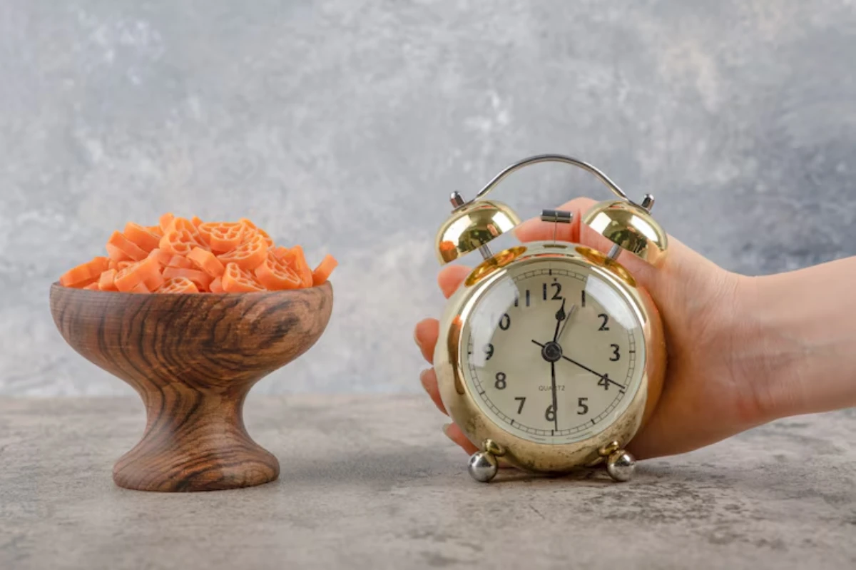 A hand holds a gold alarm clock next to a wooden bowl filled with orange pasta, set against a textured gray background.