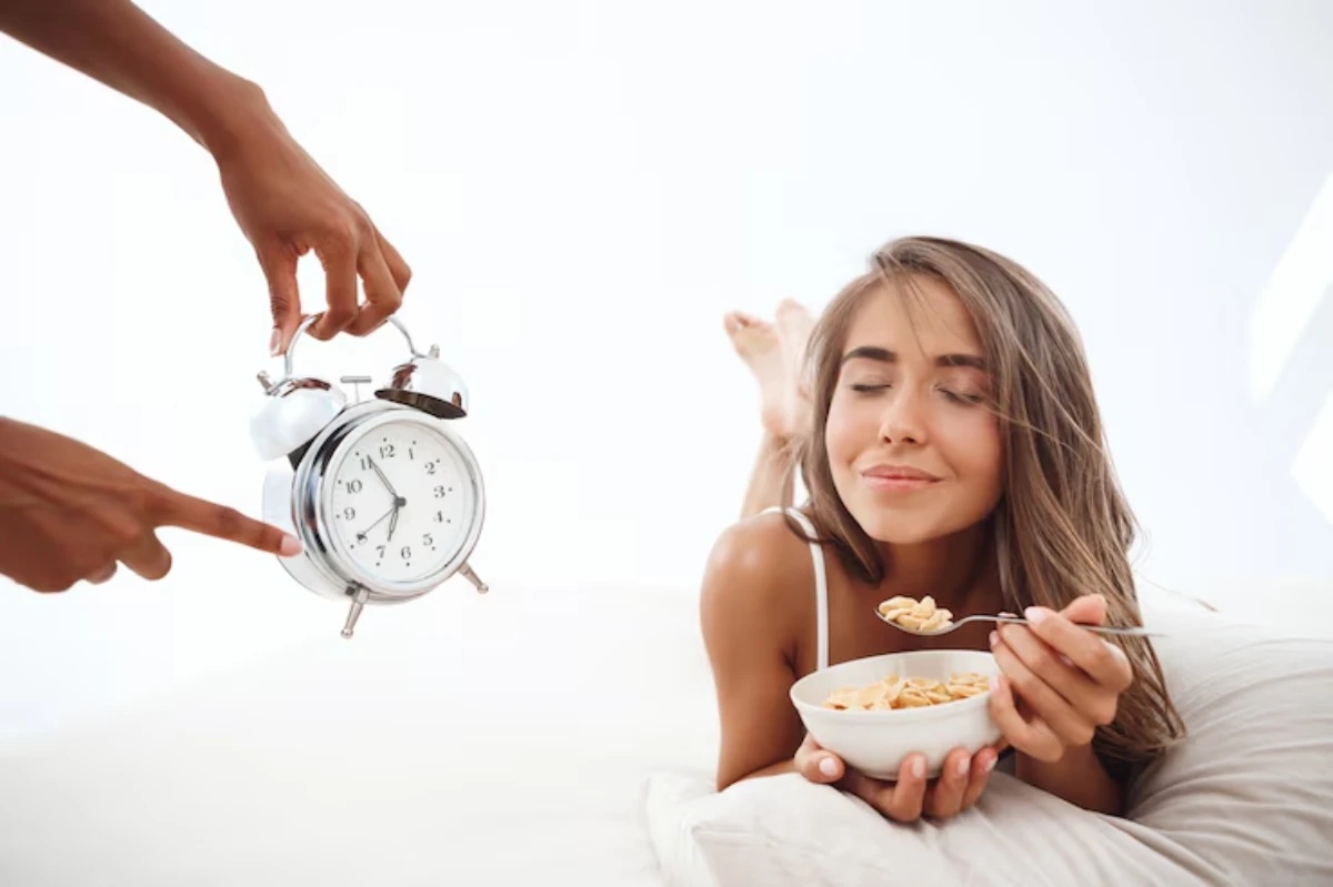 A hand holds a vintage alarm clock, while a person enjoys a bowl of cereal in bed, surrounded by soft pillows and natural light.