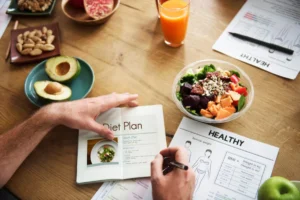 A person examines a diet plan, writing notes next to a fresh salad, fruit, nuts, and health guides on a wooden table.