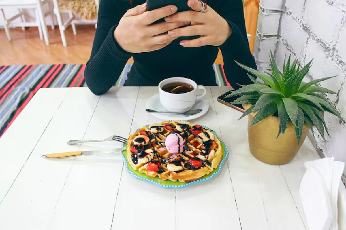 A person holds a phone above a table with a waffle topped with fruit and ice cream, a cup of coffee, and a small potted plant.