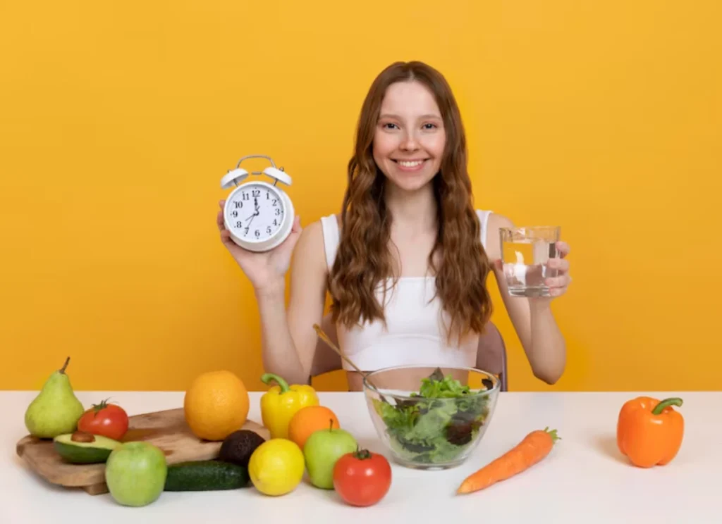 A woman holds an alarm clock and a glass of water, surrounded by fresh fruits, vegetables, and a salad bowl, against a yellow background.
