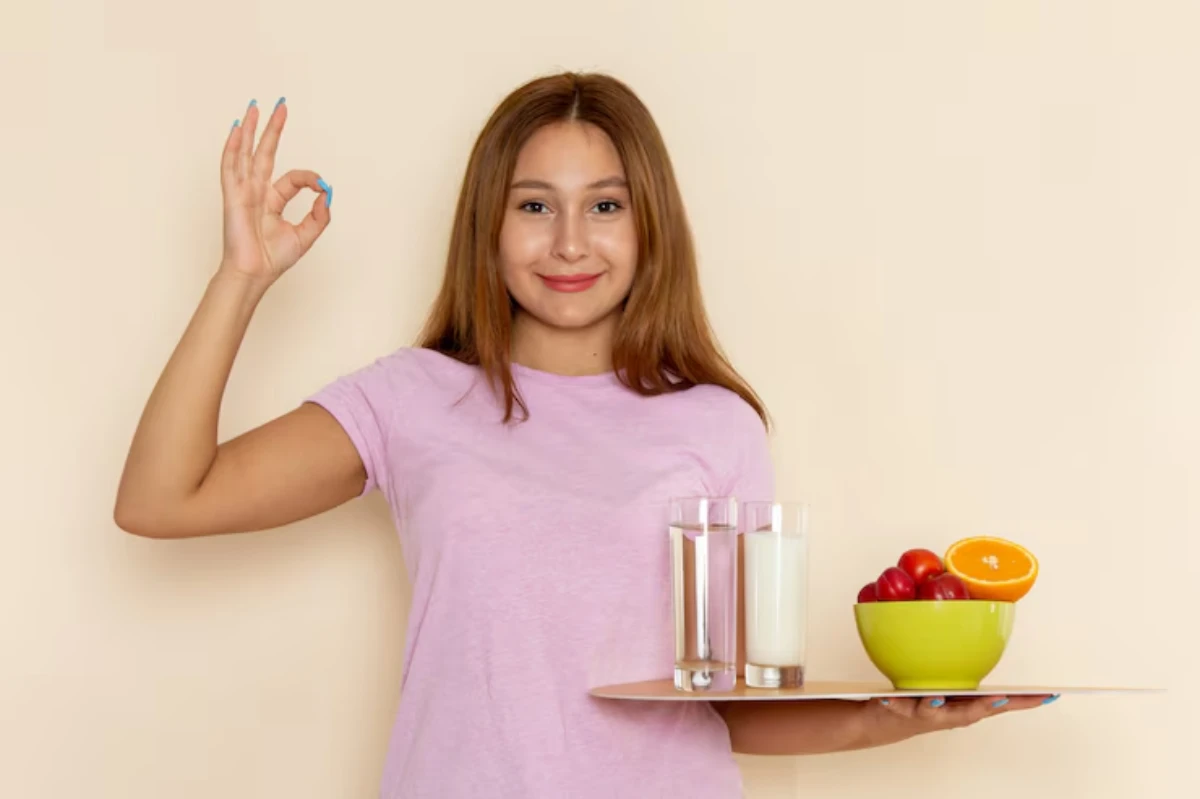 A person in a pink shirt holds a tray with two glasses of drinks and a bowl of fruit, showing an OK hand gesture.