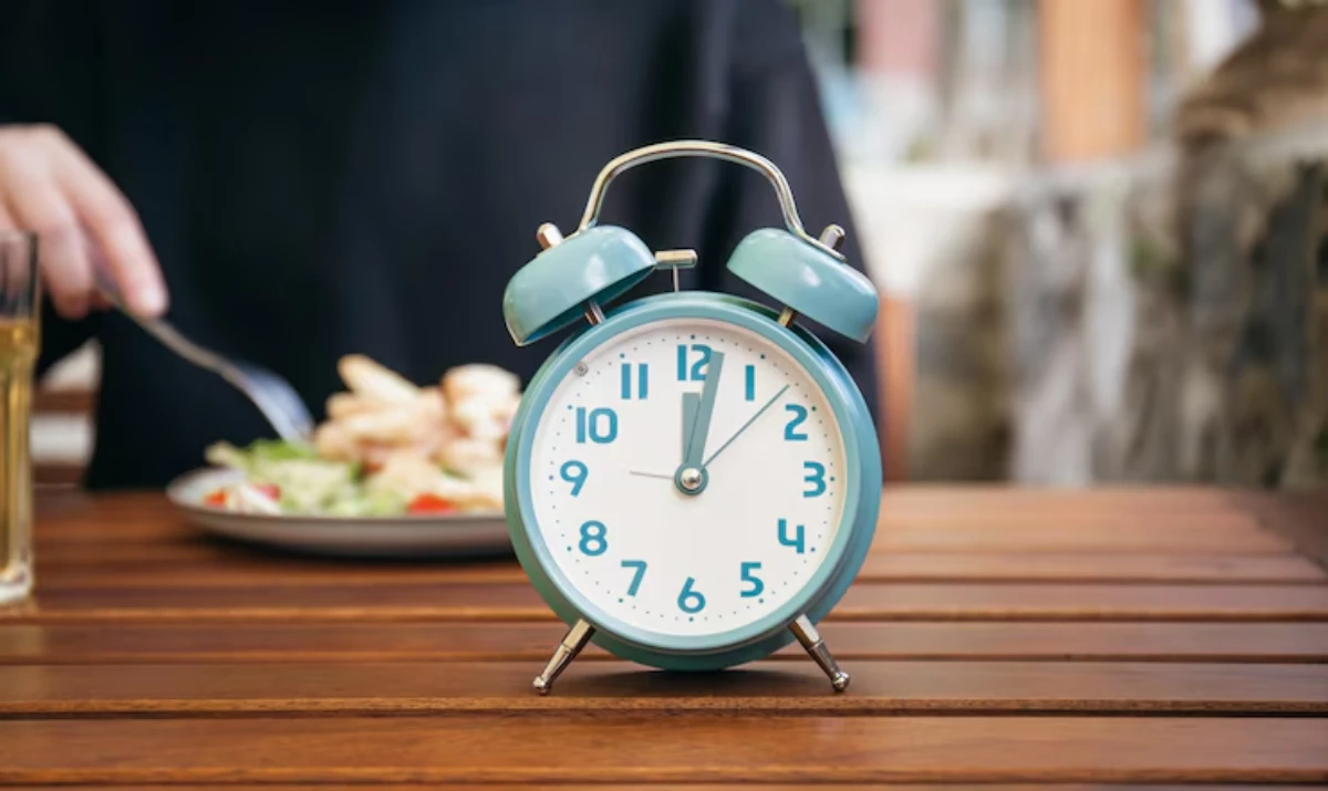 A teal alarm clock sits on a wooden table, with a plate of salad and chicken in the background, indicating mealtime readiness.