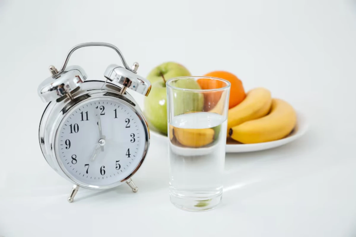 A silver alarm clock next to a glass of water, with a plate of apples, oranges, and bananas in the background on a white surface.
