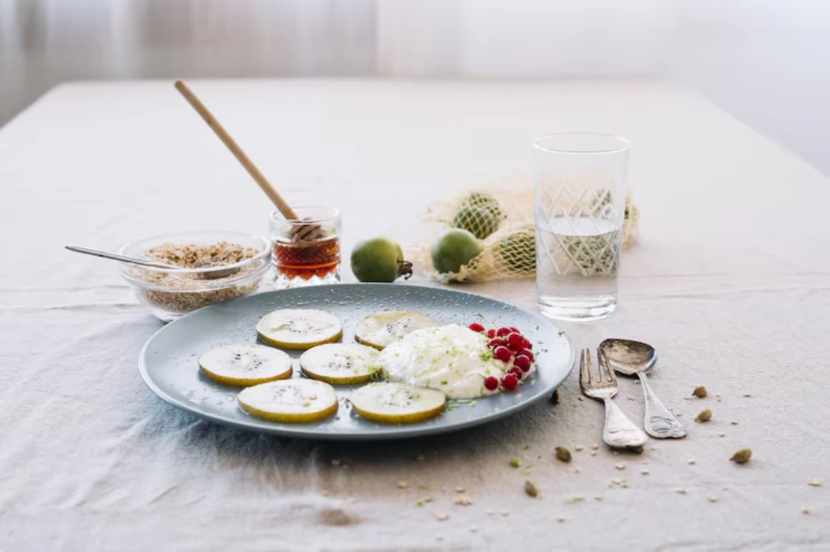 Plate with sliced pears, yogurt, and red currants, accompanied by honey, oats, a glass of water, and green apples on a table.