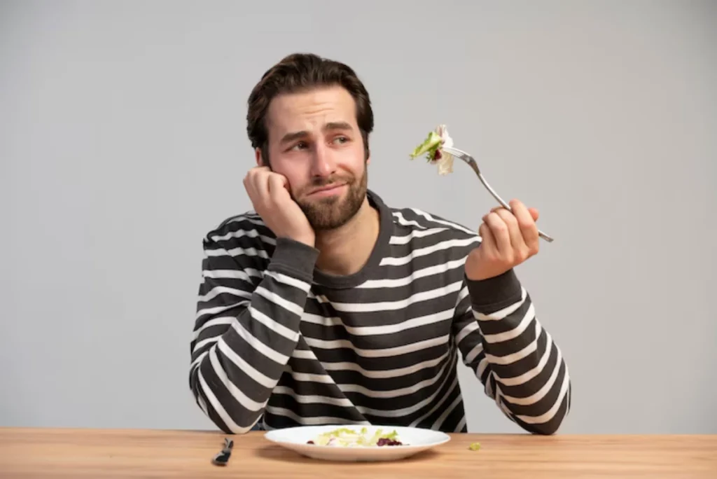 A person in a striped sweater rests their chin on their hand, holding a fork with a bite of salad, sitting at a wooden table.