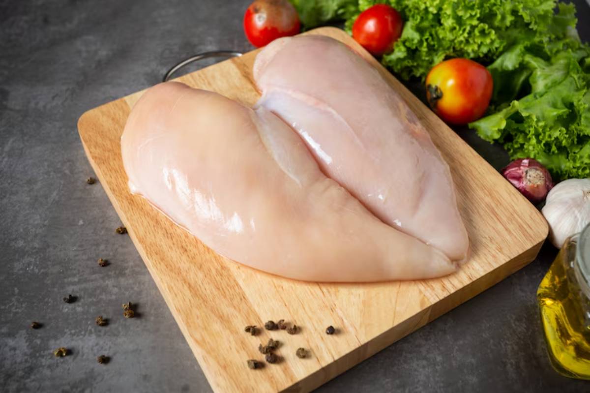 Raw chicken breasts on a wooden cutting board, surrounded by fresh tomatoes, lettuce, onions, and garlic cloves.