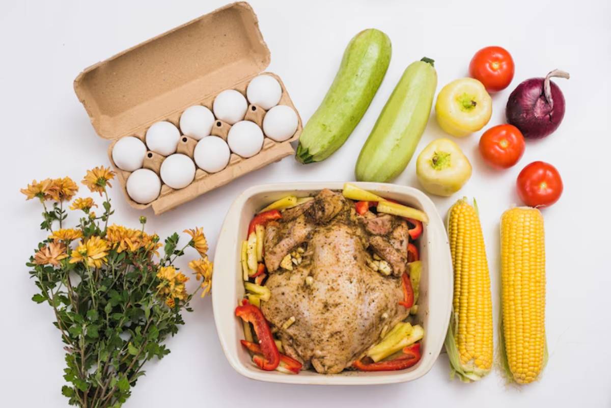 A layout of fresh ingredients: a whole chicken with vegetables, eggs, corn, tomatoes, zucchini, and flowers on a white surface.