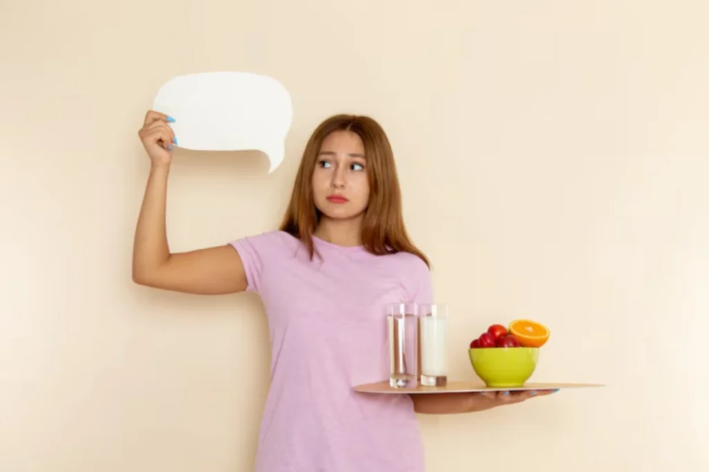 A woman holds a tray with two glasses of milk and a bowl of fruits, while raising a speech bubble in front of a light-colored background.