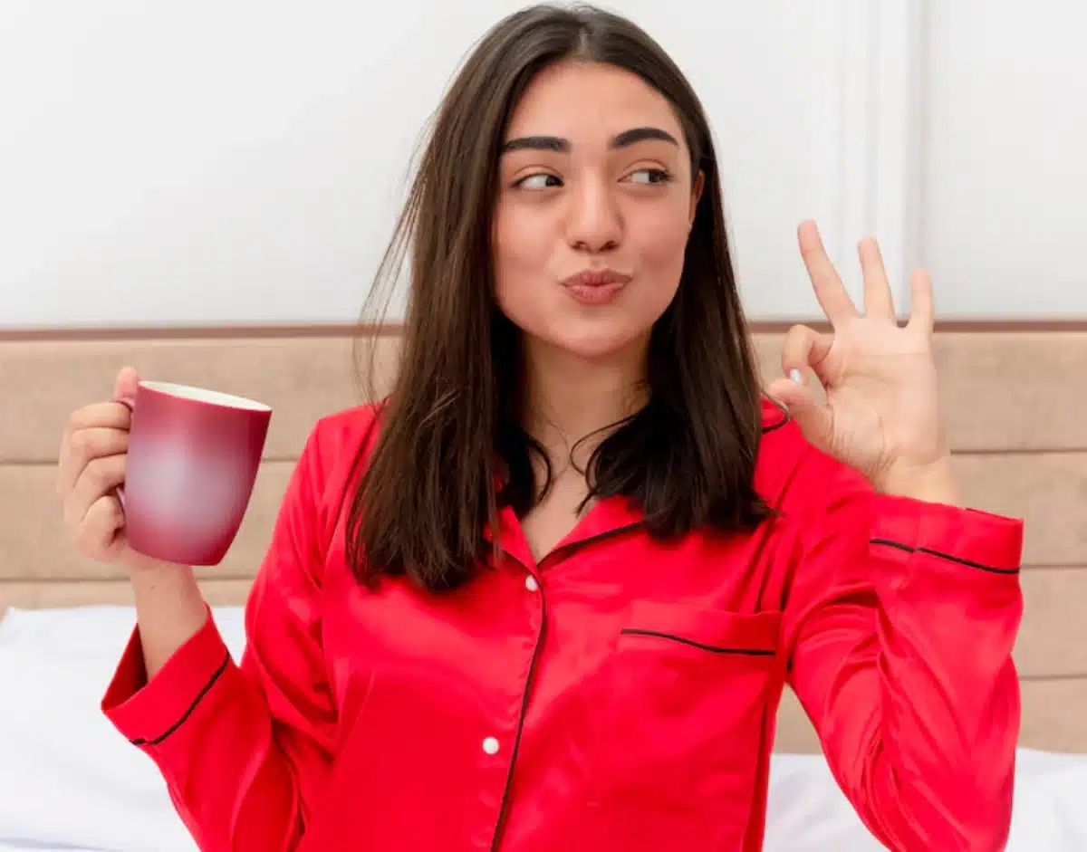 A person in a bright red pajama set holds a mug and makes an okay hand gesture, sitting on a bed with white linens.