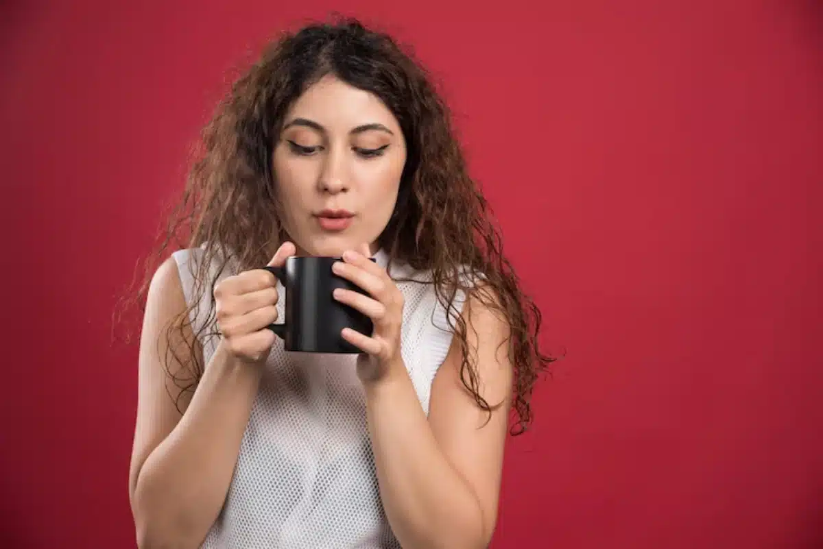 A person with curly hair holds a black mug close to their chest, set against a vibrant red background.