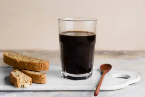 A glass of black coffee next to two pieces of biscotti on a white serving tray, accompanied by a wooden spoon.
