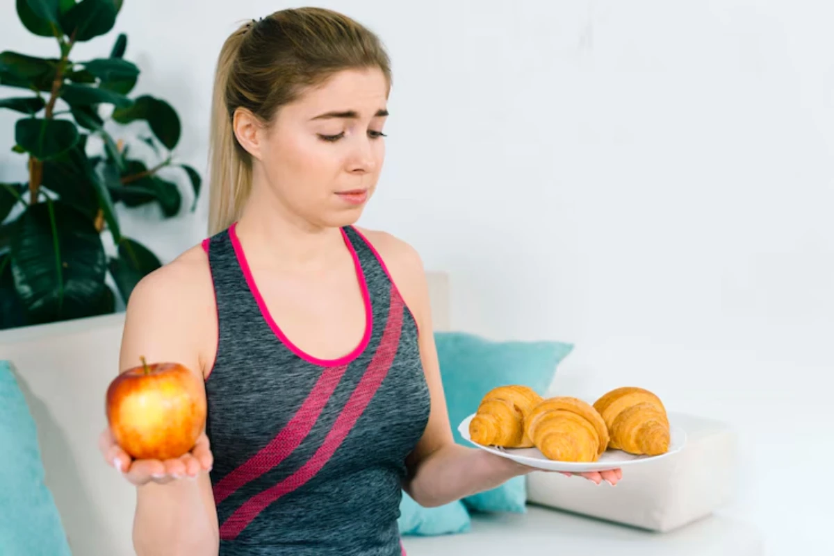 A fit woman in athletic wear holds an apple in one hand and a plate of croissants in the other, seated on a couch.