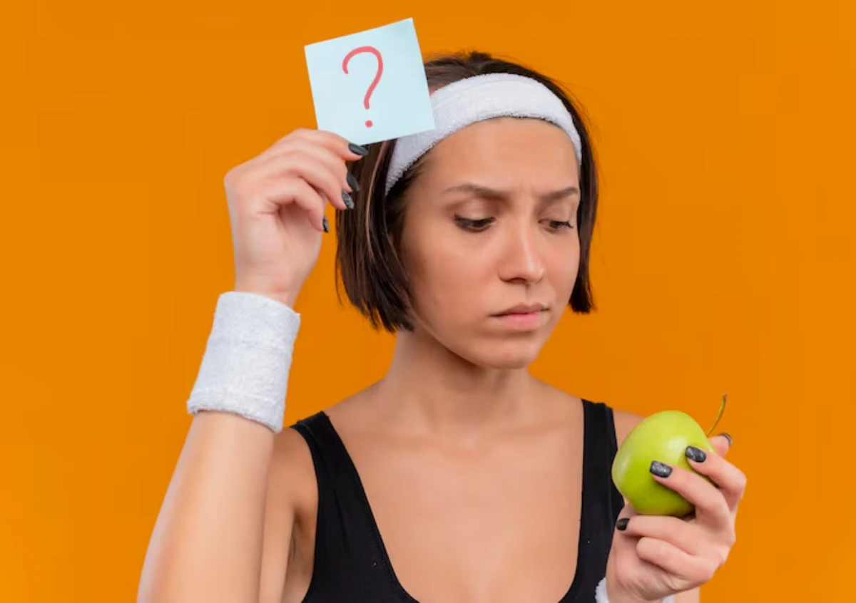 A person in a black tank top and headband holds a question mark note and a green apple against a bright orange background.