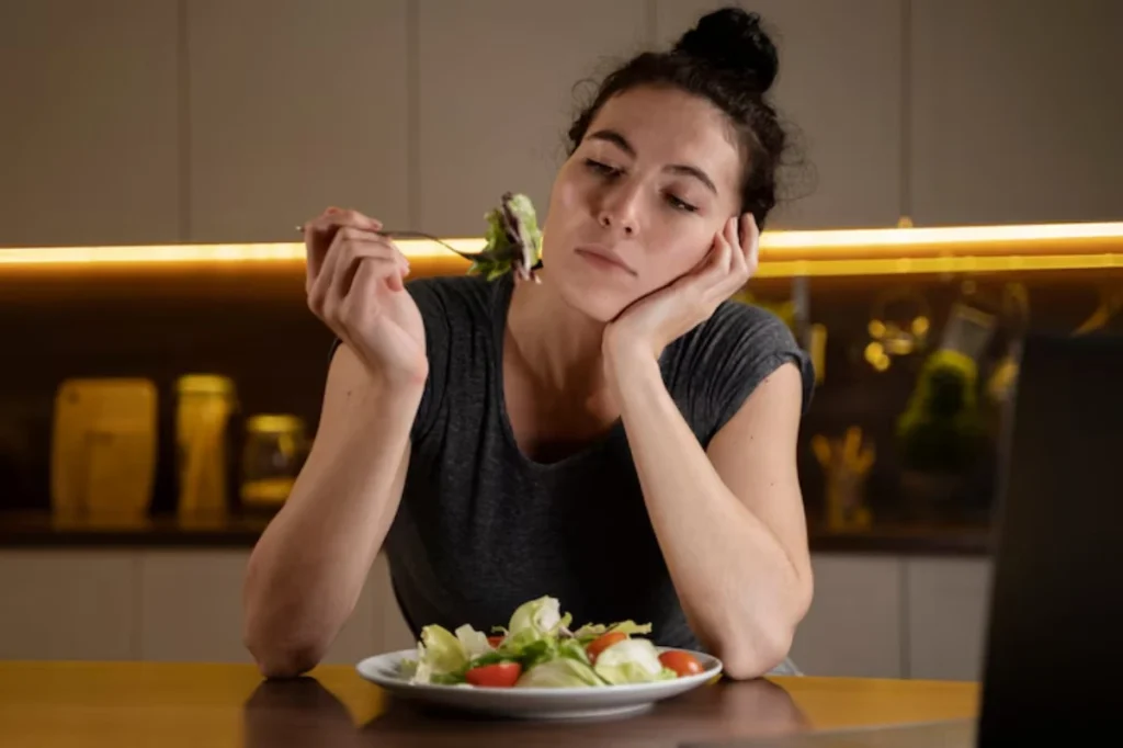 A person sits at a table, holding a fork with salad while looking thoughtfully at a plate filled with fresh vegetables.