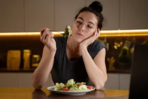 A person sits at a table, holding a fork with salad while looking thoughtfully at a plate filled with fresh vegetables.