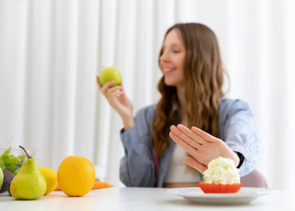 A person holds a green apple, gesturing to decline a cupcake, surrounded by various fruits on a table.