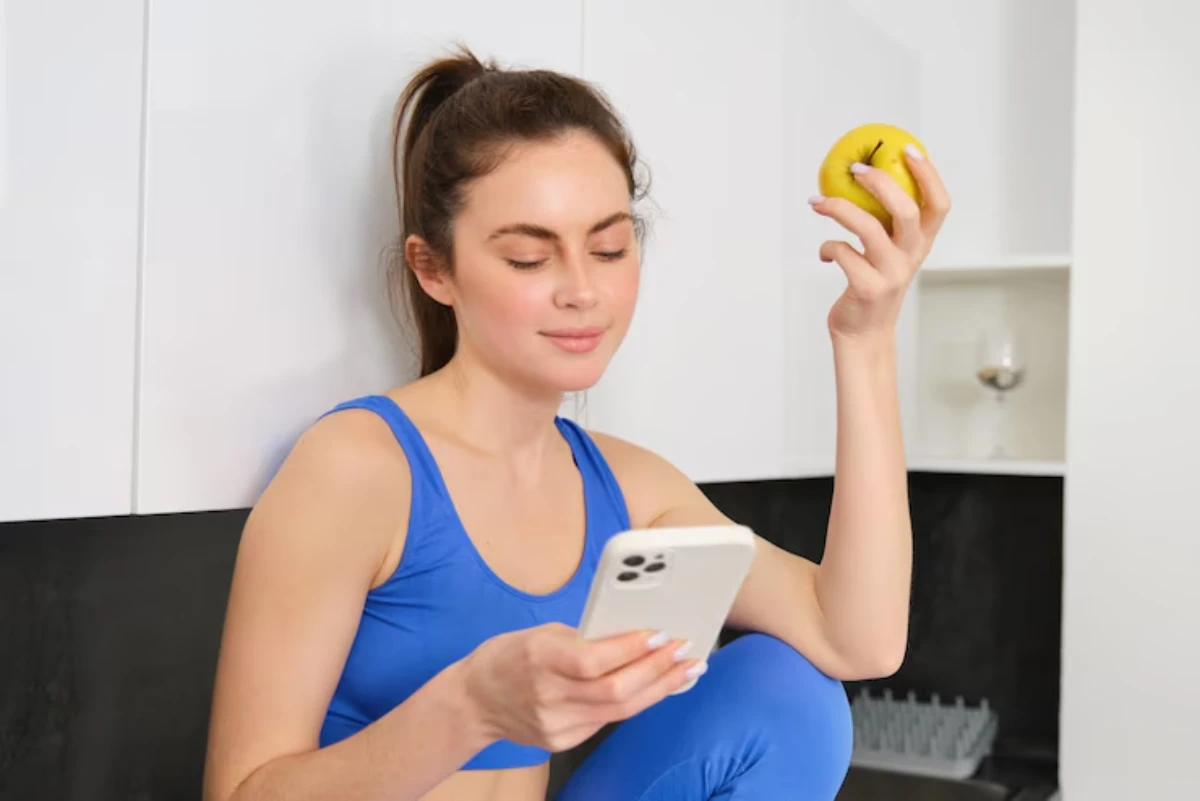 A woman in a blue workout outfit sits on a kitchen counter, holding a yellow apple in one hand and a smartphone in the other.