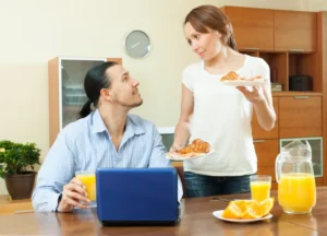 A woman serves croissants on a plate to a man at a kitchen table, with a laptop and glasses of orange juice nearby.