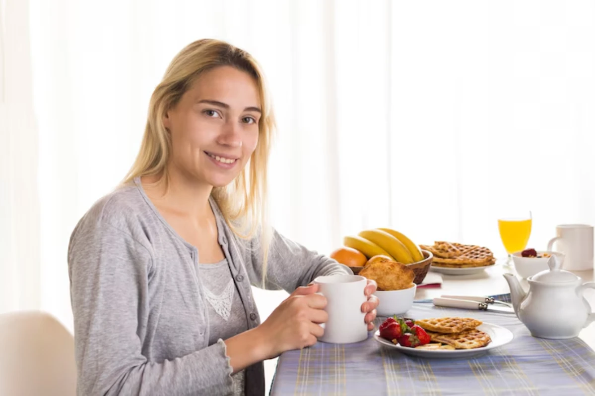 A woman in a gray cardigan sits at a table set for breakfast, enjoying coffee beside a spread of waffles, fruits, and orange juice.