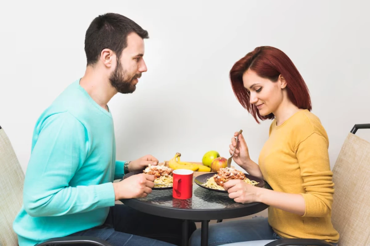 A couple sits across from each other at a table, enjoying plates of pasta and surrounded by fresh fruit and a red mug.