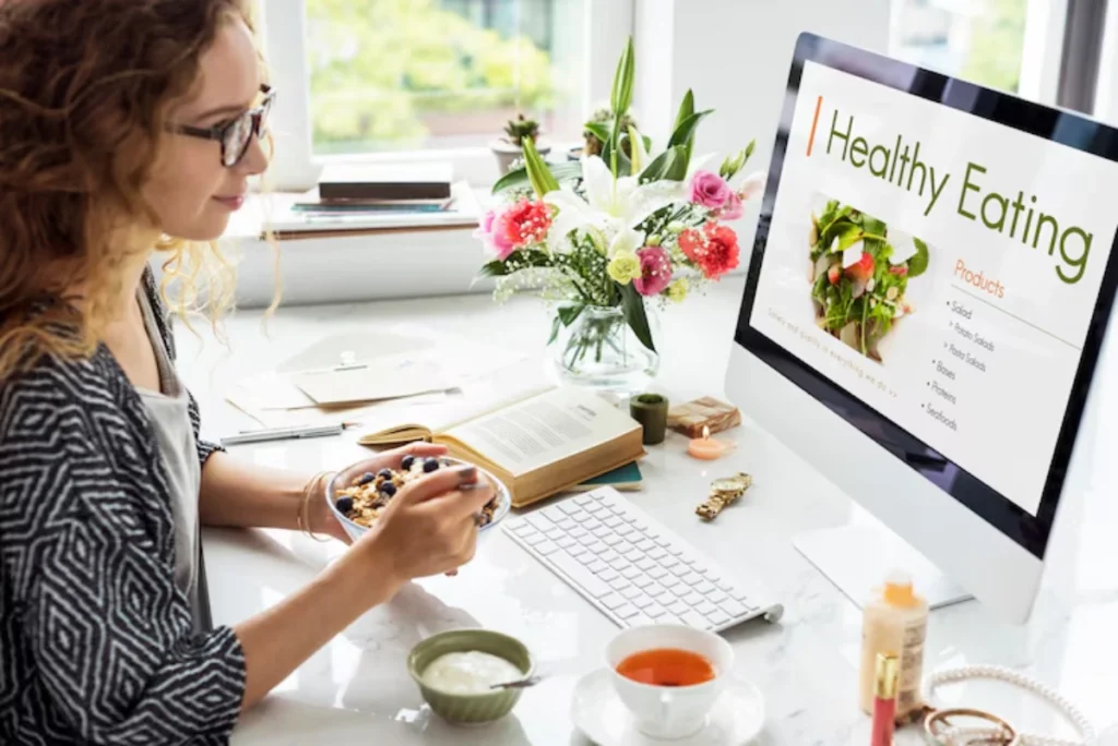 A person sits at a desk, enjoying a bowl of granola, surrounded by flowers, a book, and a computer displaying healthy eating information.