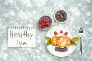 A marble plate with pancakes, fresh raspberries, and chocolate discs beside a notepad reading "Healthy Life" on a textured background.