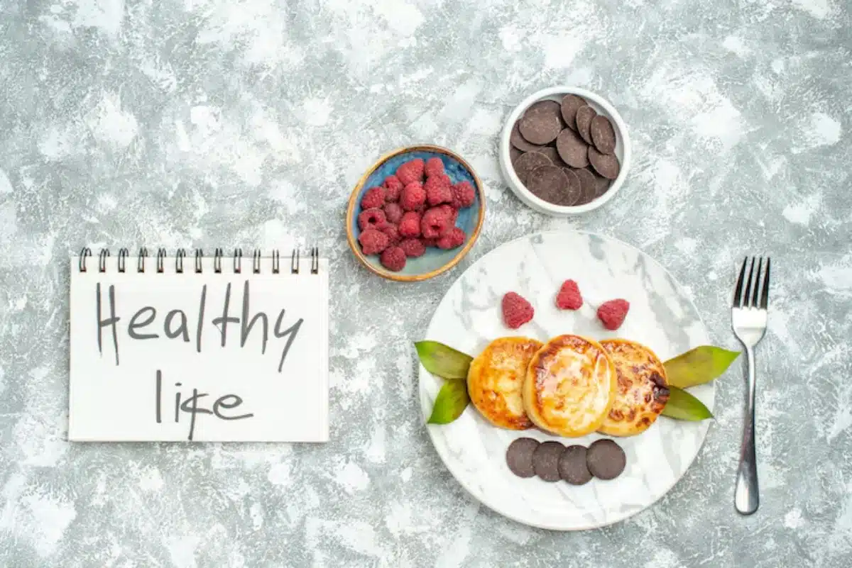 A marble plate with pancakes, fresh raspberries, and chocolate discs beside a notepad reading "Healthy Life" on a textured background.