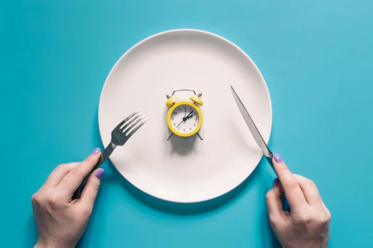 A yellow alarm clock on a white plate, flanked by a fork and knife, symbolizing mealtime and timing. Hands with purple nails are visible.