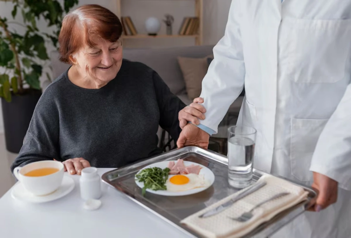 A caregiver gently assists an elderly woman at a table, where a tray with a meal and a glass of water is presented.