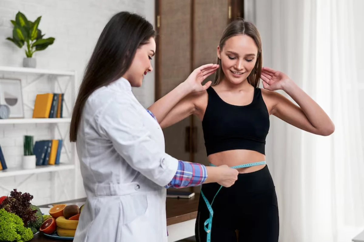 A healthcare professional measures a woman's waist with a tape measure in a well-lit, modern consultation room with plants and healthy foods.