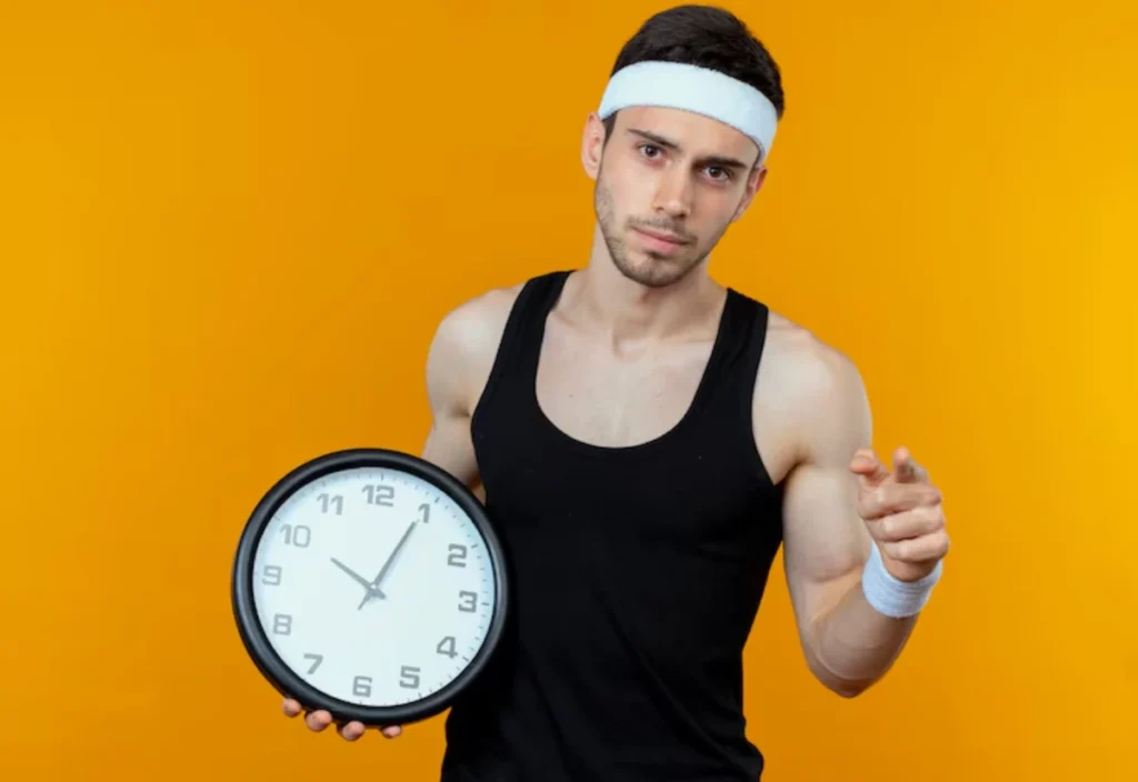 A muscular person in a sleeveless black top holds a large clock while pointing, set against a vibrant orange background.
