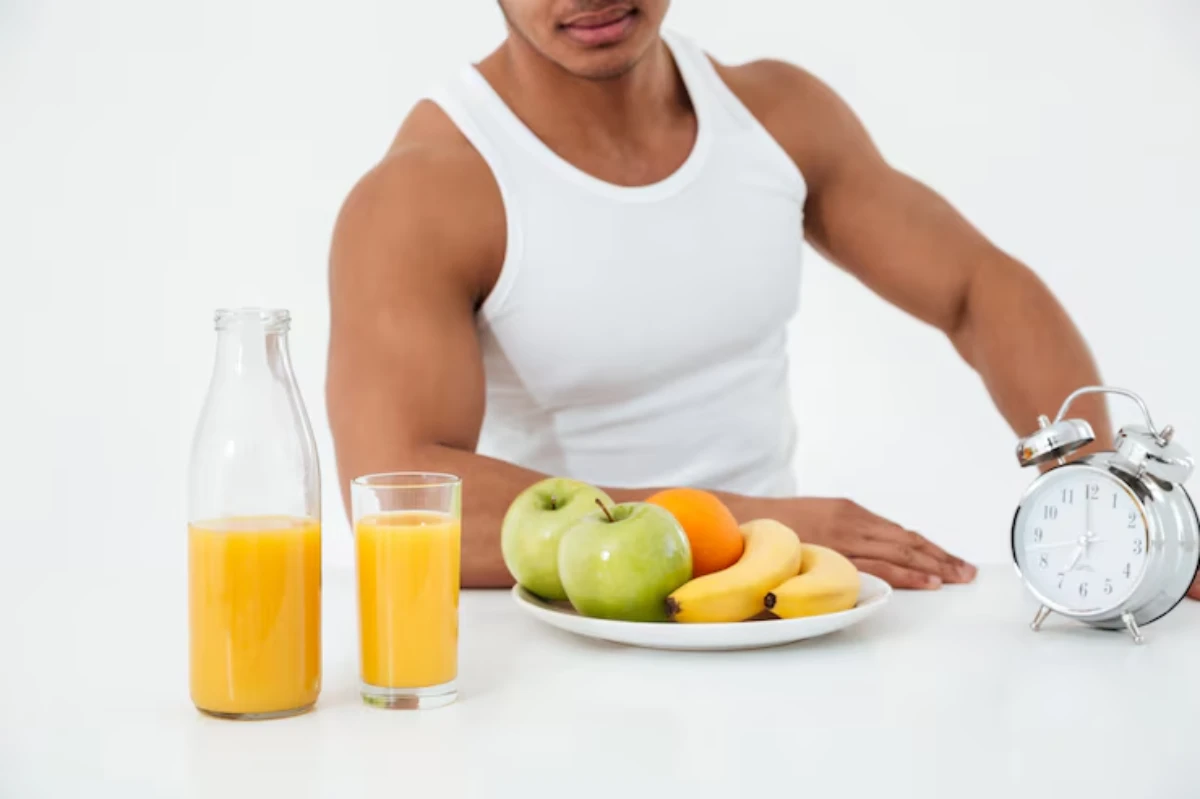 A muscular man in a tank top sits at a table with fruit, juice, and an alarm clock, promoting healthy breakfast habits.