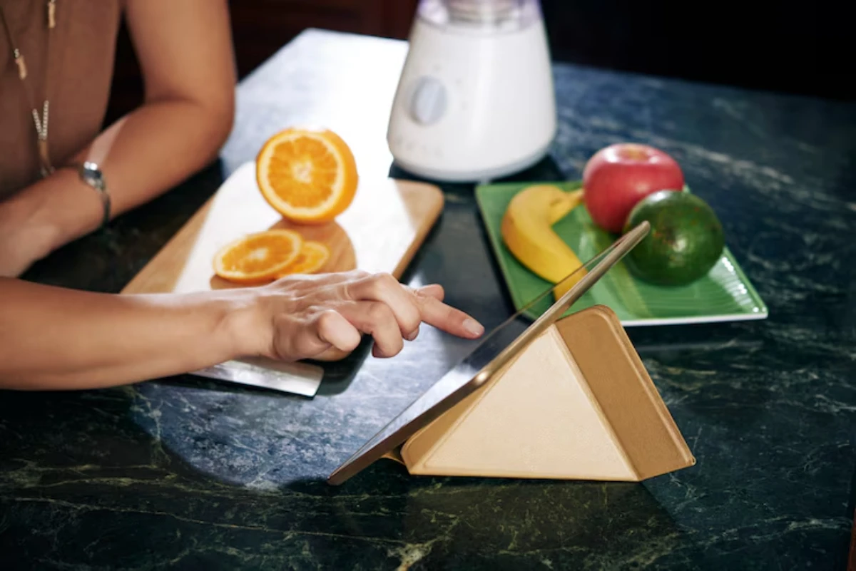 A person touches a tablet on a stand, surrounded by sliced oranges, a blender, and various fruits on green and wooden platters.