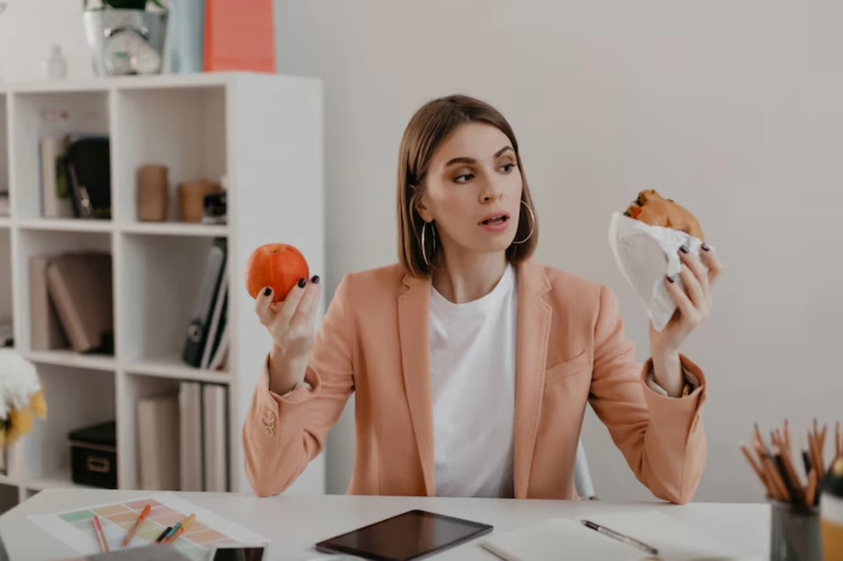 A woman in a peach blazer holds an apple in one hand and a sandwich in the other, sitting at a table with stationery and a tablet.