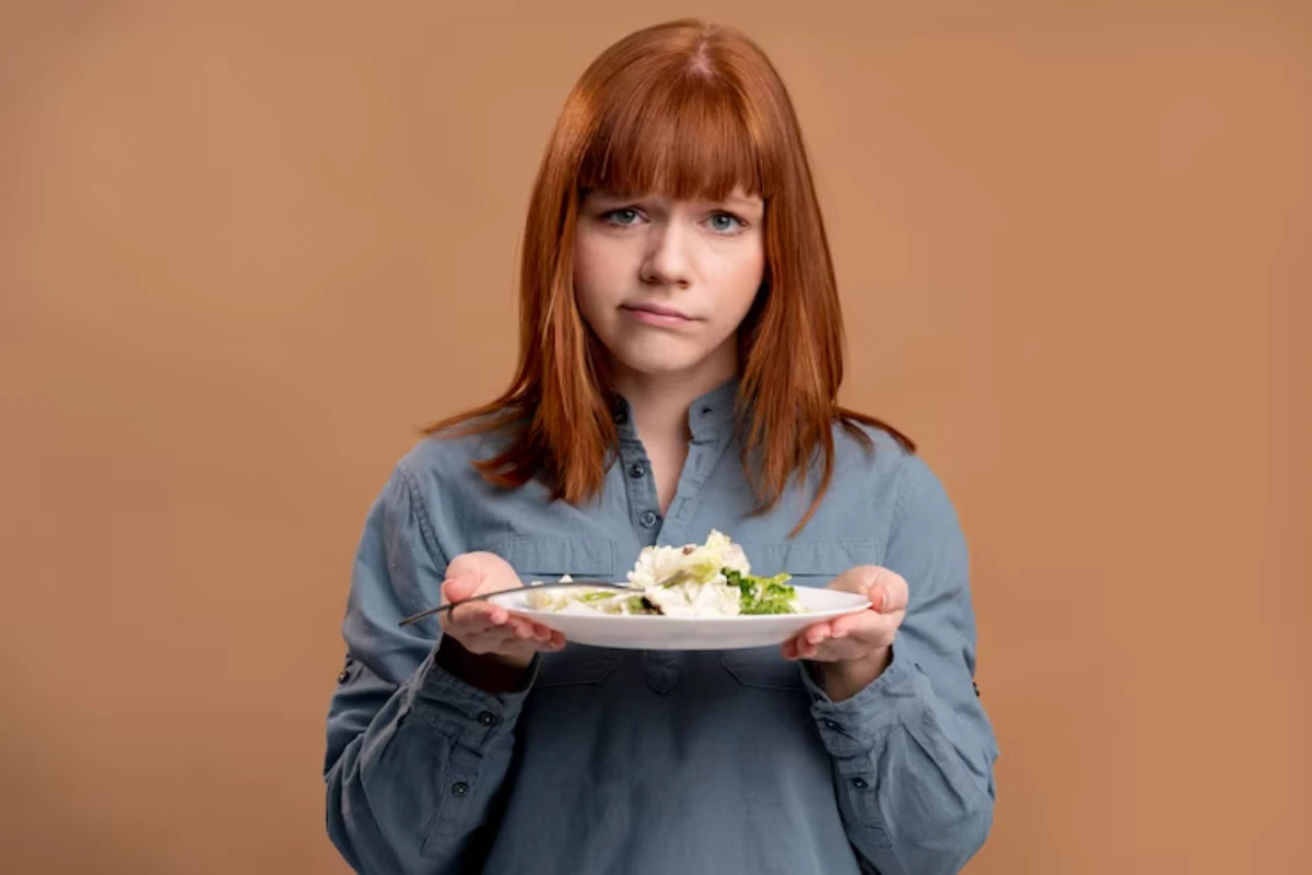 A person holds a white plate with a salad and assorted vegetables, wearing a blue shirt against a neutral background.