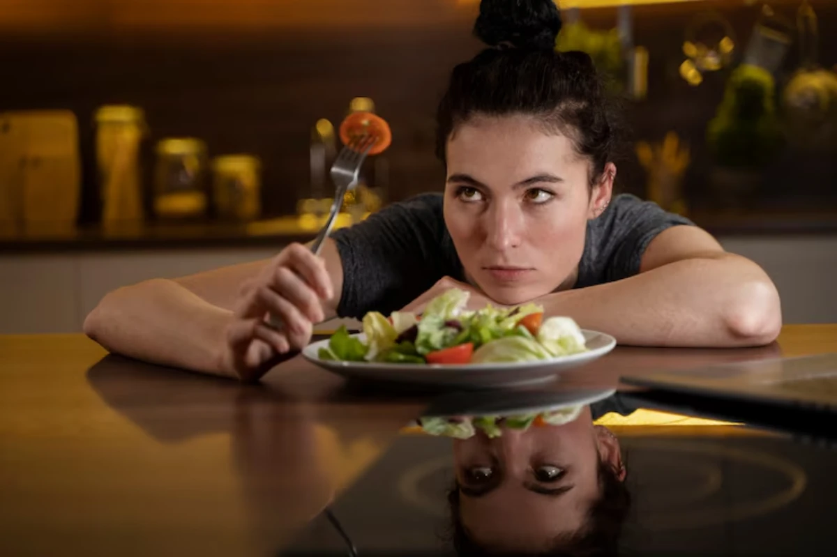 A hand holding a fork with a cherry tomato, poised above a plate of fresh salad on a wooden table in a softly-lit kitchen.