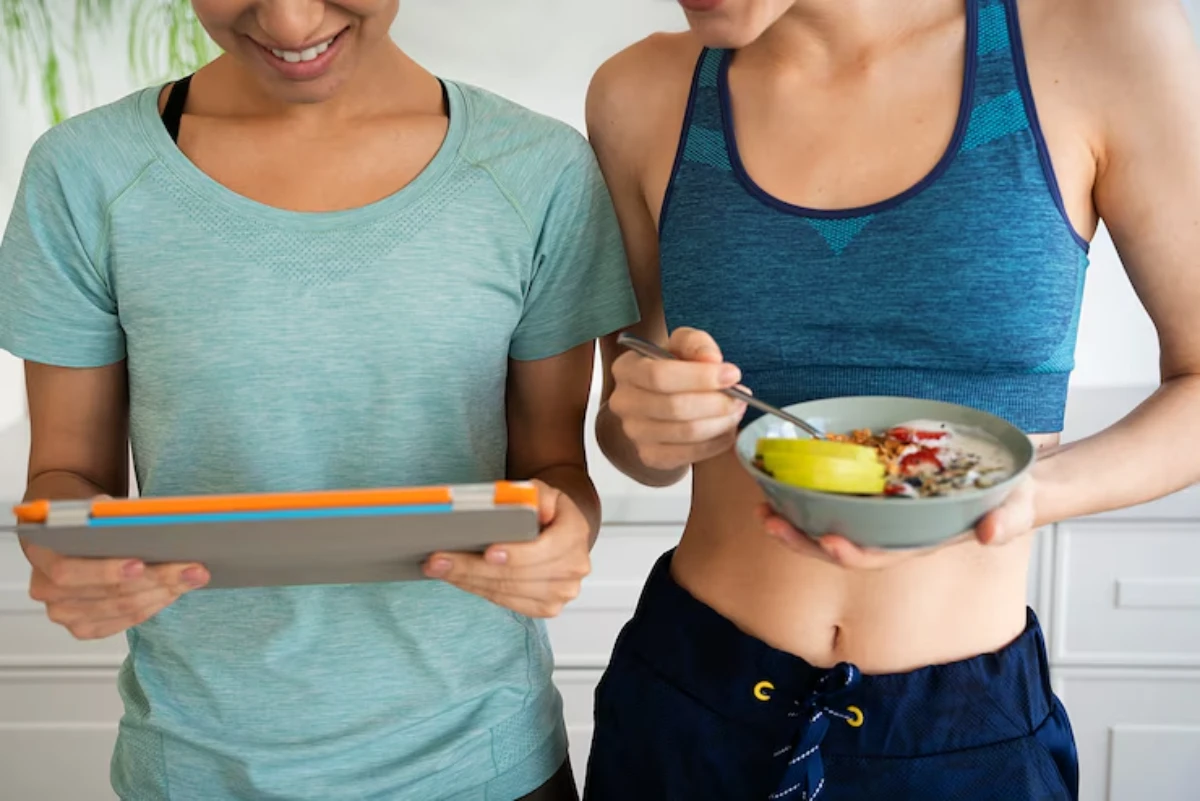 Two women in athletic wear smile at a tablet, one holding a bowl of yogurt topped with fruit, in a bright kitchen.
