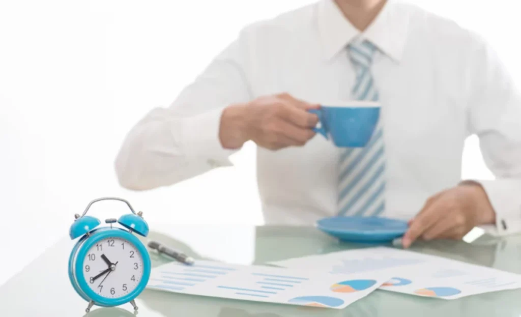 A businessman in a white shirt and tie sips from a blue cup while sitting at a desk with an alarm clock and documents.