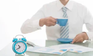 A businessman in a white shirt and tie sips from a blue cup while sitting at a desk with an alarm clock and documents.