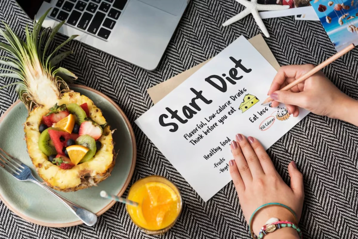 A hand holding a pencil writes on a Start Diet list near a colorful fruit bowl and a refreshing drink, on a textured table.