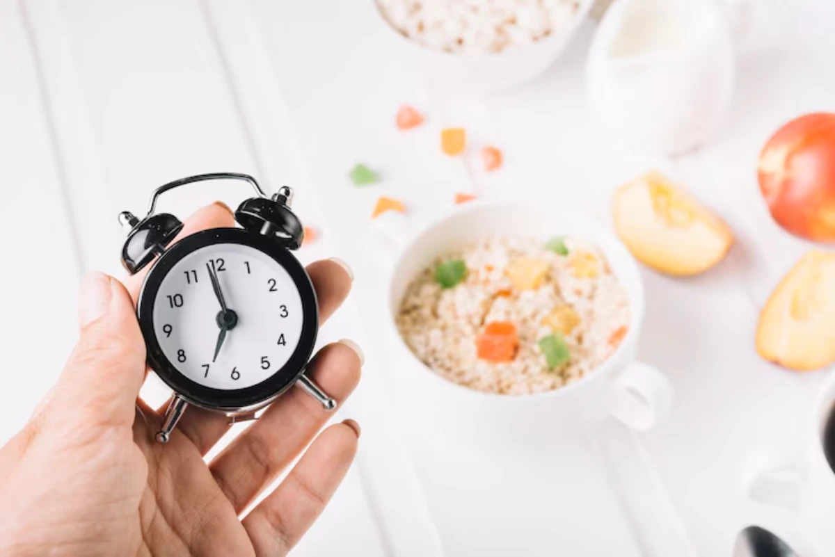 A hand holds a black alarm clock next to a bowl of colorful sprinkled cereal, sliced fruit, and a white jug in a bright setting.
