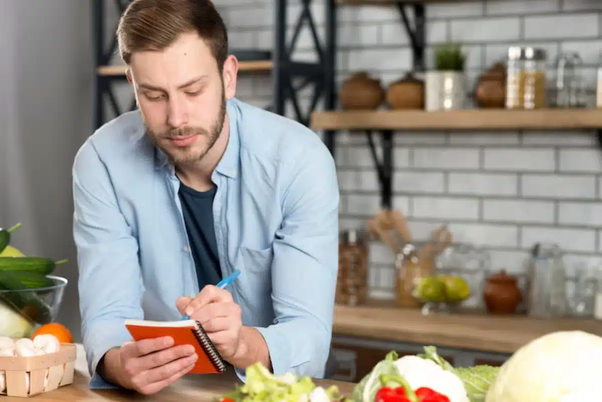 A person in a light-blue shirt writes in an orange notebook, surrounded by fresh vegetables in a bright kitchen.