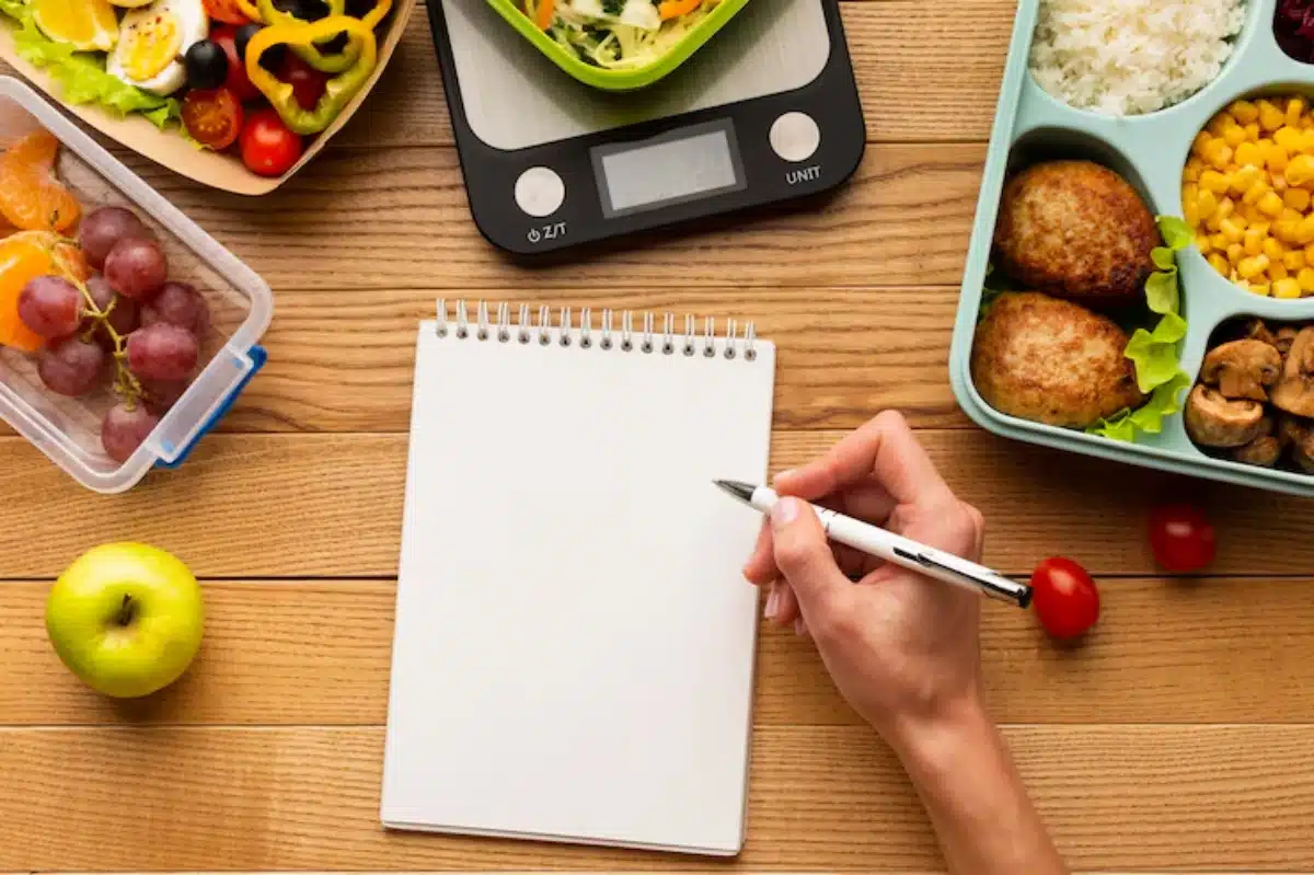 A hand holds a pen over a blank notebook, surrounded by healthy food options and a digital scale on a wooden table.