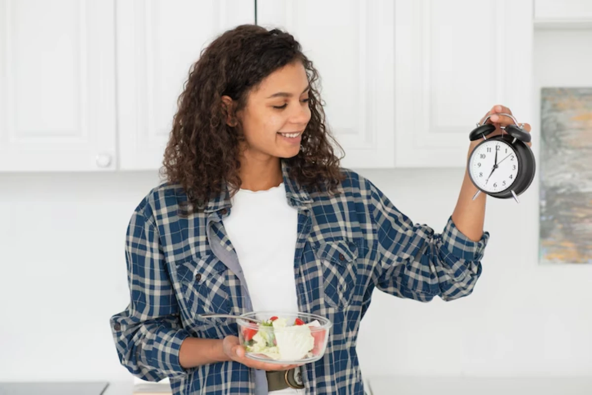 A person in a plaid shirt holds a bowl of salad in one hand and an alarm clock in the other, standing in a bright kitchen.